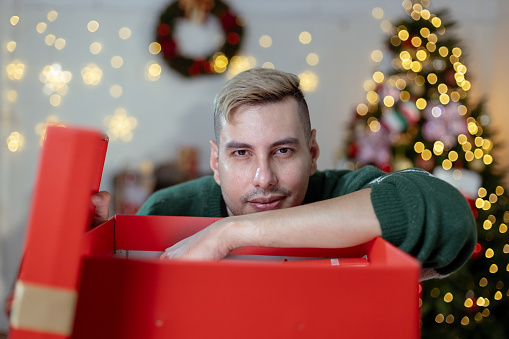 Christmas day concept. handsome man sitting surprised and opening gift box with christmas tree background. Festive atmosphere party.