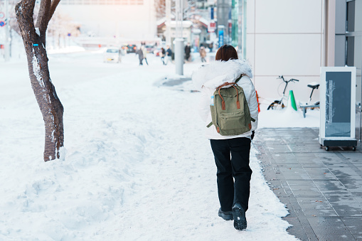 happy Traveler with Sweater and backpack walking on snow covered road in frosty weather, woman tourist sightseeing in Sapporo city, Hokkaido, Japan Winter Travel and Vacation concept