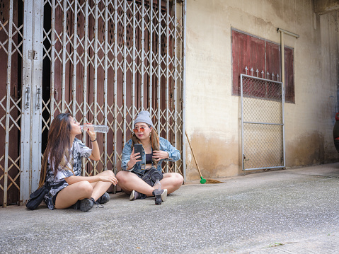 Happy two young Asian friends using mobile phone and drinking water while sitting at old folding metal gate in background, Happy friends relaxing and spending time together at outdoors, People and lifestyle