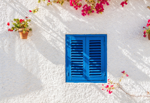 A blue wooden shuttered louvred window of a white washed spanish town house, with Bougainvillea and potted plants against the white washed wall.