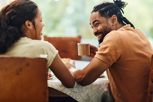 Happy African American man talking to his wife while drinking coffee during breakfast at picnic table in the backyard.