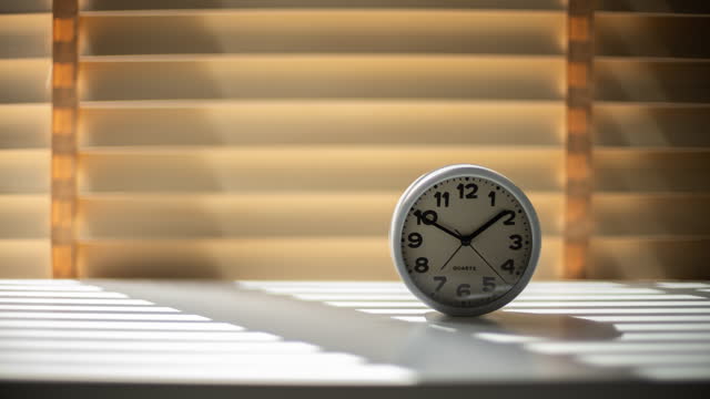 Sunlight shining through window blinds and its shadow appearing on a clock inside a house and the light is getting darker as the time passes by in the afternoon.