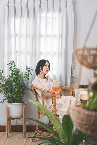 People lifestyles at living home apartment concept. Young asian woman reading a book in cozy room style with plant wood table and lamp light. Girl with short hair and casual cloth. Youth culture style