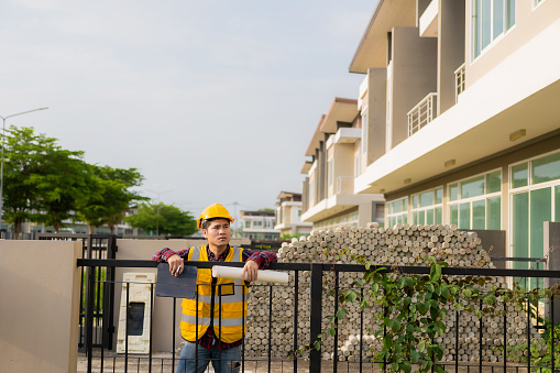 Young Asian civil engineer or construction supervisor wearing a helmet looks away and smiles while inspecting a construction site.