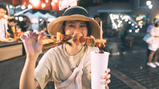 Happy young asian foodie woman eating bbq grilled skewers at outdoor night market street food vendor Poeple travel and eating street food concept. Happy young adult asian foodie woman holding bbq grilled skewers at outdoor vendor night market. night market stock pictures, royalty-free photos & images