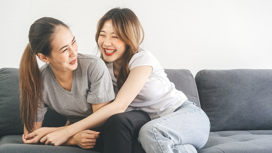 Two young adult woman living together with relationship at home concept. Southeast asian people couple funny embracing happy romantic moment on sofa.