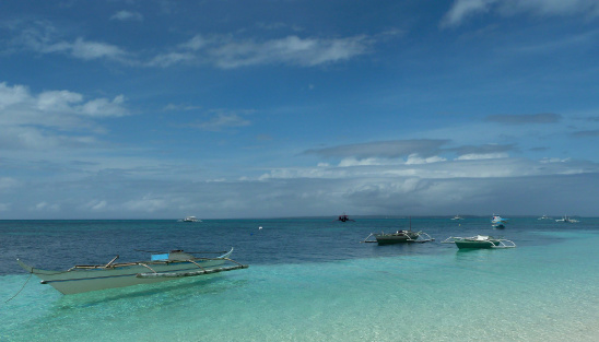 Tropical island in the Philippines on a sunny day. In the foreground, a traditional Filipino white boat takes center stage, peacefully floating in the pristine waters of a crystal clear sea lagoon. In the distance, the island reveals its scenic beauty with lush hills adorned in abundant tropical plants. The vibrant blue sky above is adorned with fluffy clouds, completing the picturesque scene.