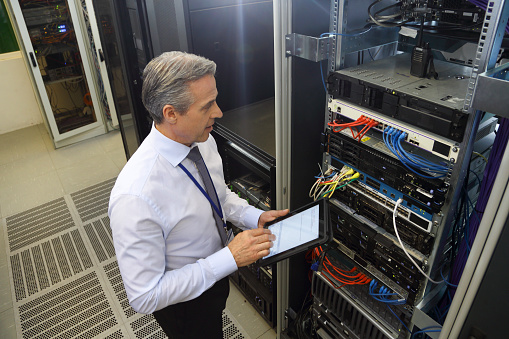 Mature adult man in data center programing on a mainframe with his tablet.
