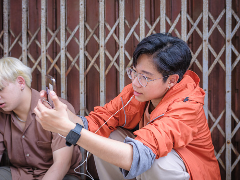 Two young Asian friends using mobile phone and sitting at old folding metal gate in background, Happy friends relaxing and spending time together at outdoors, People and lifestyle