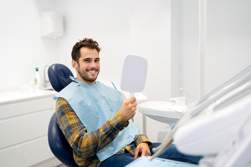 Satisfied man smiling at camera while holding a mirror after a whitening procedure in a dental clinic