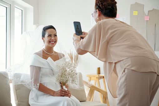 Happy bride posing for photo when mother photographing on smartphone