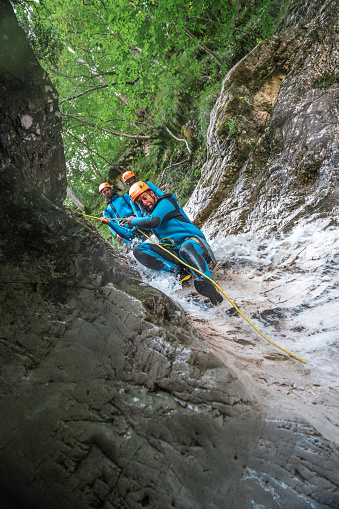 A mid-adult Hispanic male, wearing a neoprene suit and safety helmet, slides down the rugged terrain while on a canyoning adventure.