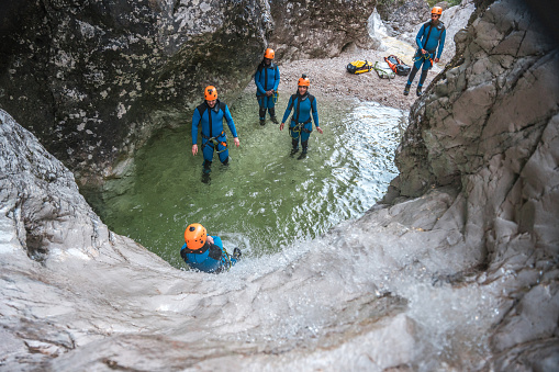 People from diverse backgrounds, in neoprene suits and safety helmets, slide down a canyon wall, having a blast during their canyoning experience.