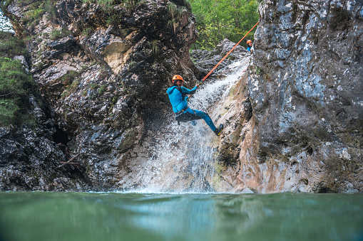 A black male in a neoprene suit and helmet rappels down a rugged canyon wall, showcasing the adrenaline-pumping adventure of canyoning.