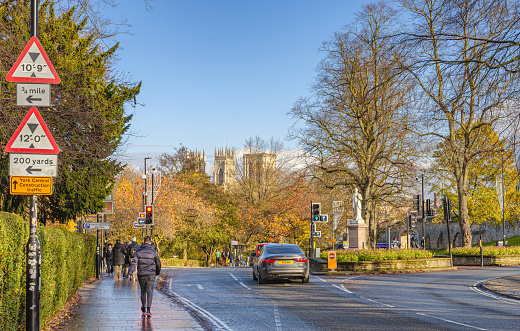 York, UK. November 17, 2023.  A city road junction with traffic lights. A statue stands near a flower bed and an ancient city wall runs down the side. The towers of York Minster rises above the trees.