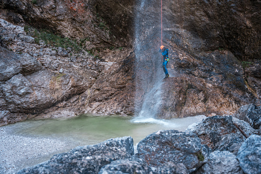 An individual, precisely geared in a neoprene suit and safety helmet, expertly descends a stunning waterfall using a secure rappel rope, showcasing the exhilaration of a canyoning adventure.