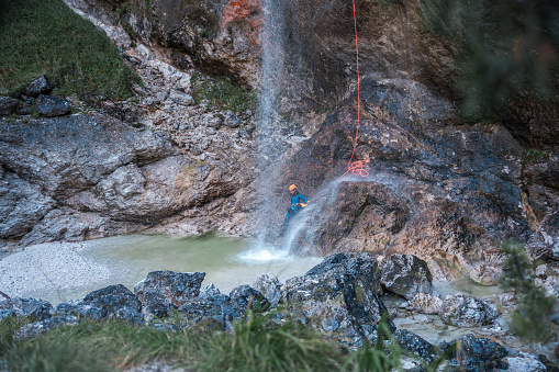 An individual in a neoprene suit and safety helmet, descending a waterfall with a rappel rope during an exciting canyoning adventure.