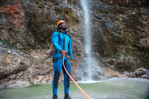 A black male in a neoprene suit and safety helmet, gears up with a rappel rope for a thrilling canyoning descent.