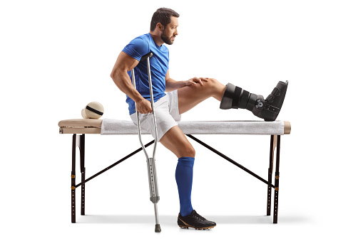 Male athlete with crutches sitting on a physical therapy bed isolated on white background