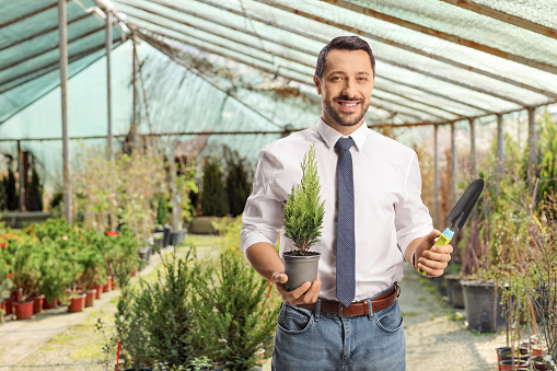 Smiling businessman holding a small tree in a pot and a spade in a garden