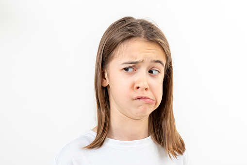 A teenage girl in a white T-shirt grimaces with displeasure on a white background isolated, doubts something, copy space.