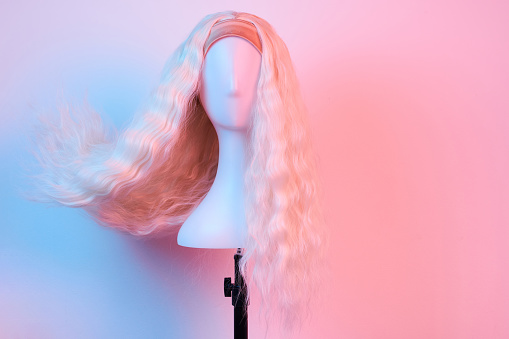 Natural looking blonde wig on white mannequin head. Long hair on the plastic wig holder isolated on white background pink lighting, front view