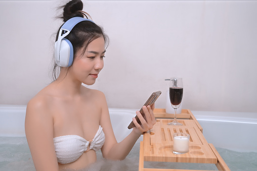 Asian woman luxuriates in the bathtub, utterly immersed in the soothing melodies, as she interacts with her smartphone, responding to messages and enjoying the digital connection.