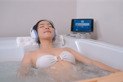 Asian woman luxuriates in the bathtub and closes her eyes, utterly immersed in the soothing melodies playing through her digital tablet, the perfect environment for relaxation.