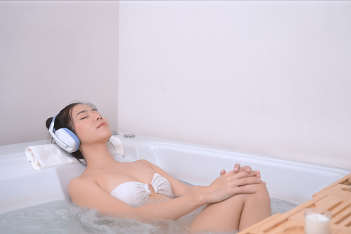 Asian woman luxuriates in the bathtub and closes her eyes, utterly immersed in the soothing melodies playing through her digital tablet, the perfect environment for relaxation.