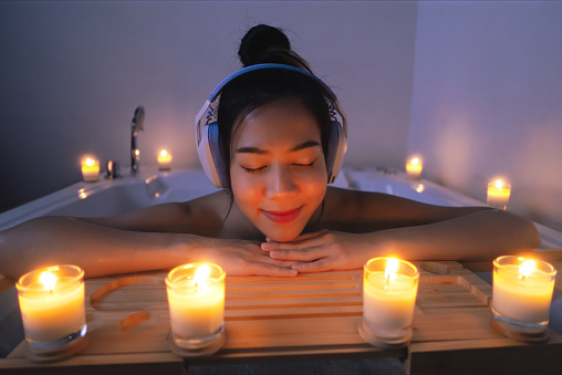 Close-up of Asian woman luxuriates in the bathtub and closes her eyes, utterly immersed in the soothing melodies. The scented candle and warm glow illuminates the bathroom, creating a cozy atmosphere.