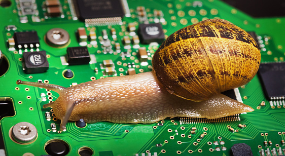 A snail on a green circuit board, part of a computer hard drive controller board. The circuit board is populated with electronic components like capacitors, resistors, a CPU, and solder points. The snail's shell is brown with stripes and patterns. The snail is moving across the circuit, leaving a trail of its slime. The contrast between the natural, organic form of the snail and the manufactured, precise components of the circuit board creates an intriguing juxtaposition.