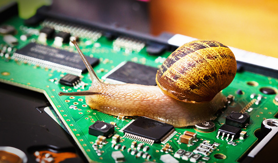 A snail on a green circuit board, part of a computer hard drive controller board. The circuit board is populated with electronic components like capacitors, resistors, a CPU, and solder points. The snail's shell is brown with stripes and patterns. The snail is moving across the circuit, leaving a trail of its slime. The contrast between the natural, organic form of the snail and the manufactured, precise components of the circuit board creates an intriguing juxtaposition.