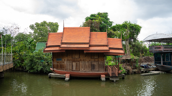 Picture of a small wooden Thai pavilion on the waterfront located on the riverbank. There is a small pier on the right side of the picture. There are trees planted at the back. Looks shady