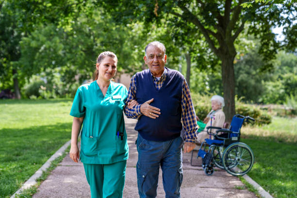 Elderly Man Assisted by Nurse in Sunny Nursing Home Garden Caring nurse supports senior man walking outdoors, with elder in wheelchair in background. aids walk stock pictures, royalty-free photos & images