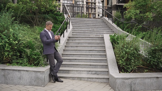 Happy businessman holding smartphone, posing near steps in the city. Business, communication, technology concept. Slow motion