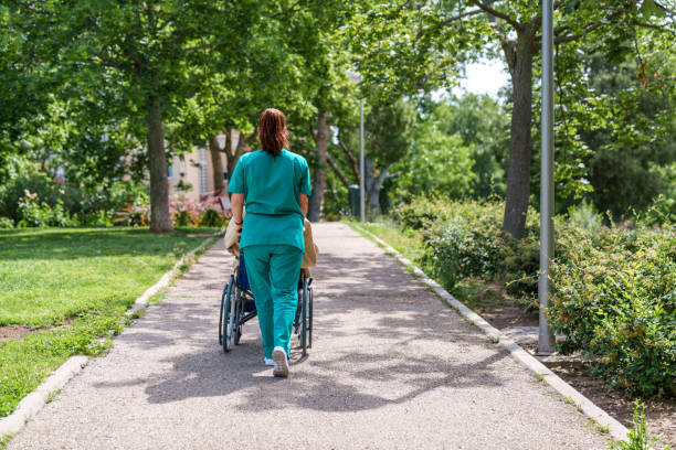 Healthcare Worker Assisting Elderly Patient in Wheelchair at Nursing Home Nurse in green scrubs pushing a wheelchair-bound senior through a garden pathway in a nursing home's yard. aids walk stock pictures, royalty-free photos & images