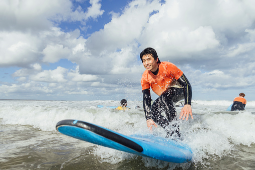 A low angle view of a young man who is taking part in a surfing lesson on the coast in Ambleside. He has taken part in a lesson with his friends and is taking to it quite well. He is looking forward confidently. He is riding a blue surfboard.