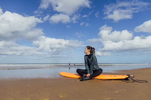A low angle view of a young woman who is taking a break from a surfing lesson with friends. She is just having a tranquil moment listen ing to the water and appreciating the surrounding as she sits on her board on the beach on Ambleside in the North East of England