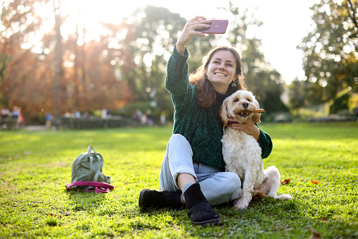 Young woman having fun taking a selfie with her soft coated wheaten terrier dog in a park. About 25 years old, Caucasian female.