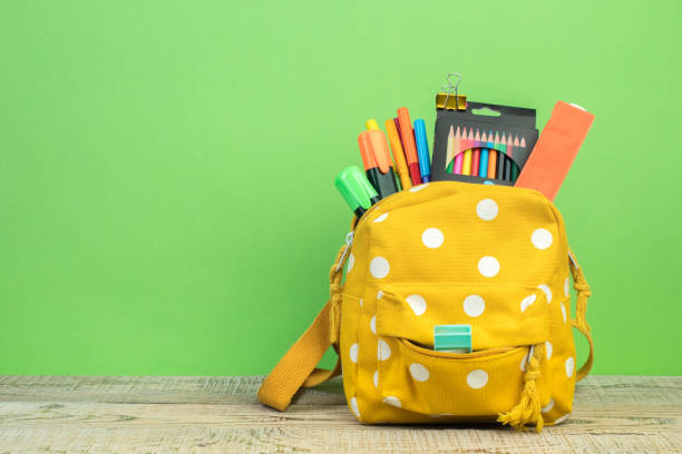 istock Yellow backpack with white polka dots with different colorful stationery on table. Bright green background. Back to school. 1816461126
