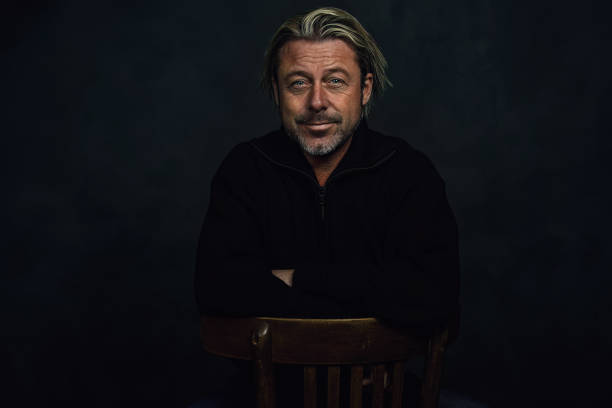 istock Smiling middle aged blond caucasian man with stubble beard in black woolen sweater sits on wooden chair in front of a dark wall. 1816462196