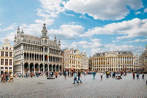 The Grand Place, a central square in Brussels, Belgium. The square is surrounded by ornate buildings, including the Town Hall and the Maison du Roi. The cobblestone square is filled with tourists and locals, and the Belgian flag is visible in the background. The sky is blue with white clouds.