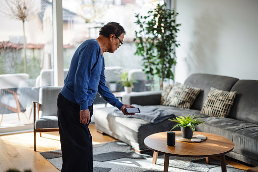 A senior adult Asian man curiously interacts with a smart speaker using his smartphone in the bright, comfortable setting of his living room.