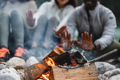 Three multiracial women are putting their hands over a bonfire to heat them, as the weather outside is cold. They are wearing jackets and their faces are unrecognizable.