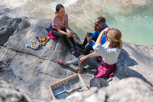 A Caucasian woman, Asian woman and a Dark-skinned man wearing sports wear are sitting together over some big gray stones while having a picnic together. They are talking and enjoying some snacks together.