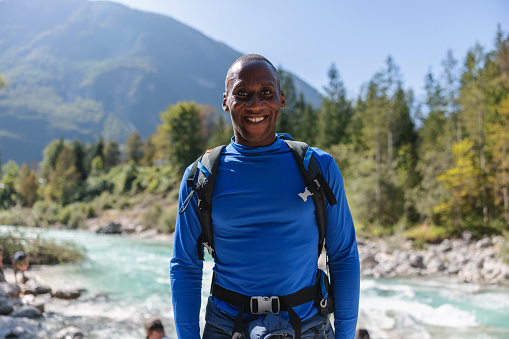 A dark skinned man wearing sports wear makes a stop during his hike at the moutains and smiles to the camera. At the back the landscape looks defocused with a river, trees, stones and a big mountain.