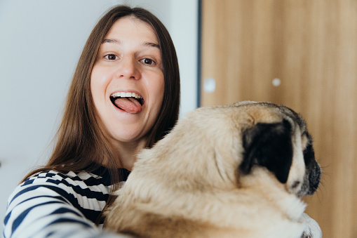 Self Portrait of smiling female with long hair, wearing striped shirt embracing with her furry pug and making photos in bright well-designed apartment during sunny day