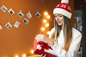 Female portrait of a beautiful woman with a Christmas cap indoors. The person is unboxing a gift box with ribbon. New Year photography, yellow background.