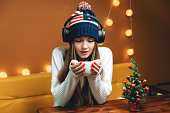 Female portrait of woman who wear american cap and wireless headphones. Christmas tree on the table.  Lifestyle photography with drink, New year photo with beautiful people indoors.