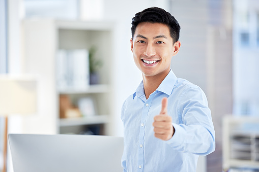Thumbs up, sign and finger being shown by a happy, smiling and pleased businessman while standing in an office at work. One young, satisfied and confident corporate professional making a hand gesture